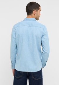 Jasnoniebieska koszula z dżinsu z yoke z tyłu, mankietami na guziki i zaokrąglonym dołem. Materiał ma lekko chropowatą fakturę i jest miękki w dotyku.