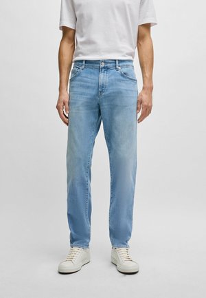 Man draagt lichtblauwe jeans met rechte pijpen, witte sneakers en een wit, kort mouw textuurtshirt, staand tegen een effen achtergrond.
