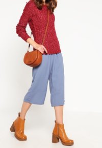 Pull tricot rouge texturé, pantalon large cropped bleu clair, et bottines à talons marron. Un sac bandoulière marron complète le look.