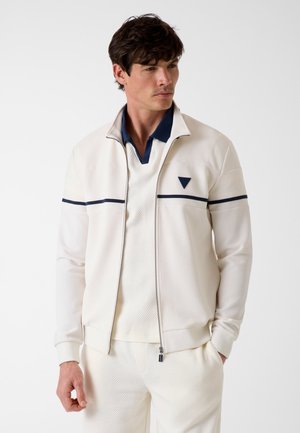 Mann trägt weiße, strukturierte Jacke mit Reißverschluss und passende Hose mit marineblauem Kragen und Streifendetails, schaut seitlich vor einem weißen Hintergrund.