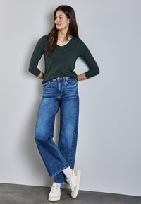 Tumma vihreä pitkähiha-paita yhdistettynä leveälahkeisiin sinisiin farkkuihin ja valkoisiin tennareihin. Kankaan vaikutelma on pehmeä ja rento istuvuus.