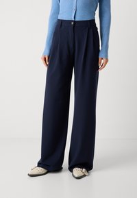 Marinefarbene weite Hose mit Vorderfalten, entworfen mit hoher Taille und einem dekorativen Knopf, kombiniert mit einem hellblauen Rippenshirt und beigen Schuhen.