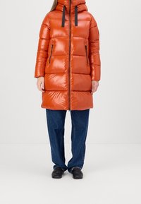 Padded orange coat with a glossy finish, featuring a high collar, black zipper, and two side pockets. Paired with loose blue denim pants.