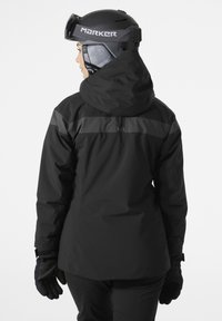 Černá voděodolná lyžařská bunda s kapucí, na ramenou ozdobena šedým horizontálním pruhem. Nosí se s černými rukavicemi, bez viditelných vzorů.