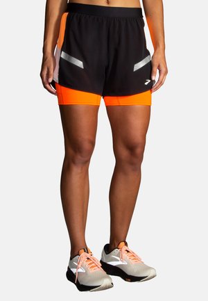 Pantaloni sportivi neri e arancioni con accenti riflettenti; il design a strati include tasche in rete. Indossati con scarpe da corsa grigio chiaro.