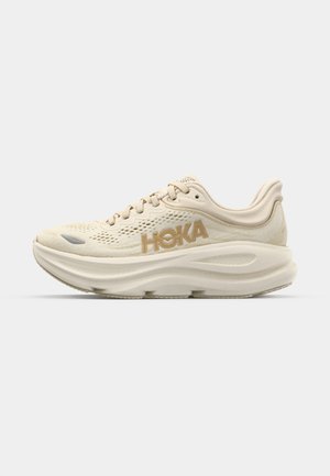 Light beige running shoe with mesh upper, padded collar, prominent "HOKA" logo in gold, and thick, cushioned sole for comfort.