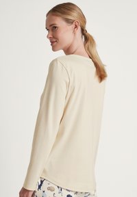Top a maniche lunghe di colore crema realizzato in tessuto morbido, con scollo rotondo e vestibilità rilassata, abbinato a pantaloni del pigiama a fantasia.