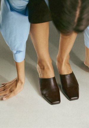 Personne portant des chaussures noires à bout carré sans lacets et un pantalon bleu, se penche en avant les mains touchant le sol sur une surface claire.
