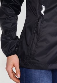 Gros plan d'une personne portant une veste noire avec une poche zippée et des poignets élastiques, associée à un jean bleu.