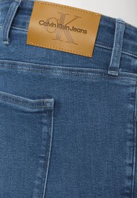 Niebieskie dżinsy z denimu mają skórzaną metkę w kolorze brązowym z wytłoczonym napisem "Calvin Klein Jeans". Materiał jest teksturowany i ma klasyczny krój.