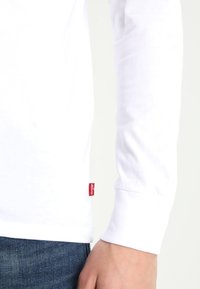 Weißes Langarmhemd aus Baumwolle mit einem roten Levi's-Logo-Tag auf der linken unteren Seite, kombiniert mit dunkelblauen Jeans.