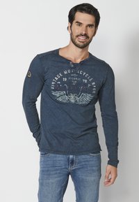 Námořnická košile s dlouhým rukávem a henley výstřihem, zdobená grafickým potiskem, který zahrnuje křídla a vintage text. Měkká, texturovaná tkanina.