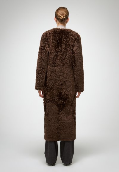 Lange, bruine jas van nepbont met een zachte textuur, oversized silhouet en rechte mouwen, gecombineerd met grijze broek met brede pijpen.