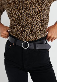 Crni kožni remen s okruglom srebrnom kopčom, nosi se preko topa s uzorkom leopard, u kombinaciji s crnim trapericama koje imaju bakrene detalje.