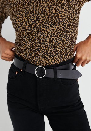 Ceinture en cuir noire avec une boucle en argent circulaire, portée par-dessus un haut en imprimé léopard, assortie à un jean noir avec des accents cuivrés.