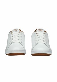 Bílé šněrovací tenisky s koženým svrškem, kulatou špičkou a gumovou podrážkou, která má hnědé akcenty. Logo viditelné na jazyku.