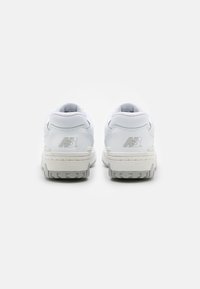 Fehér New Balance sportcipők texturált bőrből készült felső résszel, párnázott bokarésszel és szürke gumitalppal. A saroknál domború márkajelzés található.
