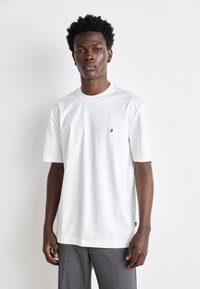 Tricou alb din bumbac cu mâneci scurte, decolteu rotund, patch mic cu logo pe piept, croi lejer, asortat cu pantaloni gri.