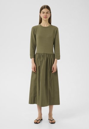Abito verde oliva con top aderente e gonna a ruota con pieghe. Maniche lunghe, tessuto morbido e orlo a ginocchio. Design semplice, senza fantasie.