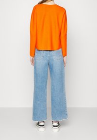 Orange Cropped-Pullover, hellblaue Schlagjeans und schwarz-weiße Sneakers. Glatte Stricktextur mit Rundhalsausschnitt und ohne sichtbare Hardware.