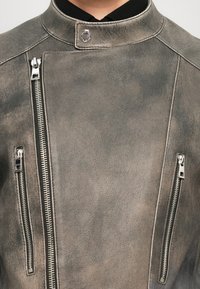 Harmaa nahkatakki, jossa on korkea kaulus, kaksi vetoketjullista etutaskua ja metalliset yksityiskohdat. Tasainen pinta ja kuluneen näköinen viimeistely.