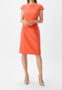 Robe corail à manches courtes avec une silhouette ajustée, coupe au genou et détails de coutures subtils. Texture lisse. Portée avec des talons nus.