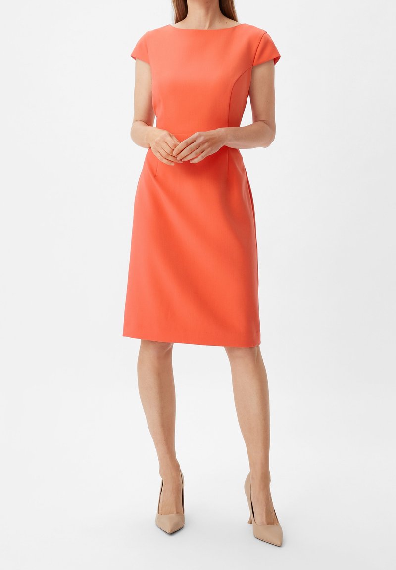 Robe corail à manches courtes avec une silhouette ajustée, coupe au genou et détails de coutures subtils. Texture lisse. Portée avec des talons nus.
