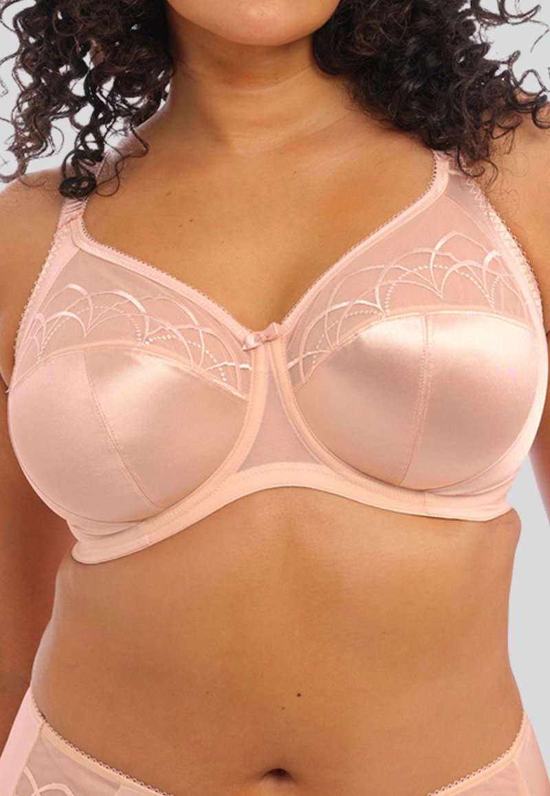 Donna con capelli ricci che indossa un reggiseno di satin e rete rosa chiaro con dettagli ricamati e una coordinata di lingerie.