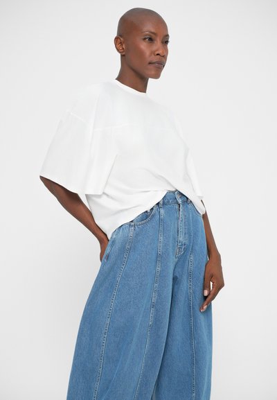 Vrouw met een geschoren hoofd, gekleed in een losse witte T-shirt in haar brede, hoge taille blauwe jeans, staand tegen een effen achtergrond.
