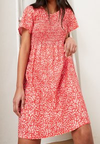 Červené květinové šaty s krátkými rukávy, které mají smocked živůtek a mírně rozšířenou sukni. Látka má lehký, texturovaný vzhled.