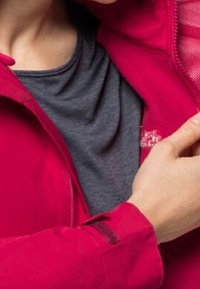 Červená bunda na zip s detaily ze síťoviny, nošená přes šedé tričko. Má boční zip a logo na bundě. Celkově hladká textura.