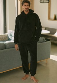 Fekete bolyhos loungewear szett, amely kapucnis pulóverből áll, elülső zsebbel és laza szabású nadrággal, elasztikus mandzsettákkal, textúrált felülettel.