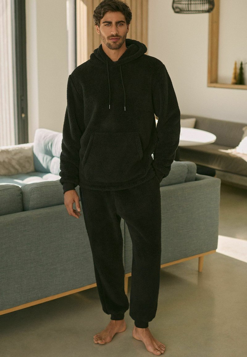 Fekete bolyhos loungewear szett, amely kapucnis pulóverből áll, elülső zsebbel és laza szabású nadrággal, elasztikus mandzsettákkal, textúrált felülettel.