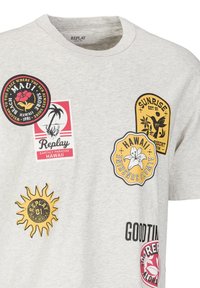 Lysegrå t-shirt med forskellige farverige Hawaiianske patches, herunder blomsterdesigns og sloganer relateret til strandliv og paradis.