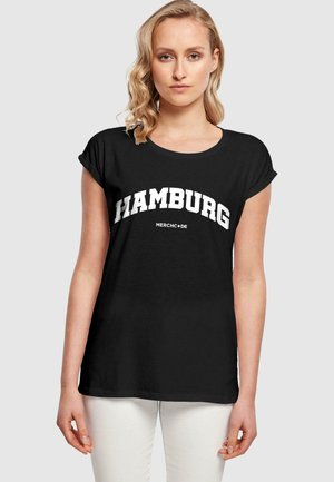 Juoda marškinėliai su surištomis rankovėmis, ant kurių yra balta išlenkta "HAMBURG" spauda ir mažesnis tekstas "MERCH•DE" po ja. Kasdienis modelis, pagamintas iš medvilnės.