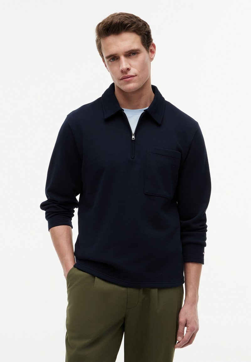 Jeune homme portant un pull bleu marine foncé avec un col zippé et une poche avant, associé à un pantalon vert olive, debout avec une main dans la poche.