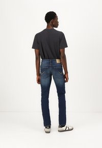 Jean en denim bleu avec une coupe droite, dotés d'une patte en cuir beige et de coutures jaunes sur les poches. Associés à un t-shirt sombre et des baskets.