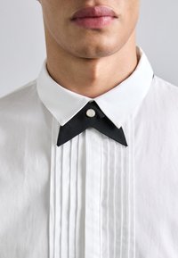White shirt with vertical pleats; features a contrasting black collar with pointed ends and a silver button detail at the collar's center.