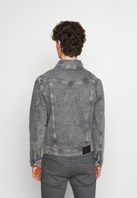 Giacca di jeans grigia con colletto classico, polsini con bottoni e dettagli di cucitura, mostrata da dietro con un'etichetta nera.