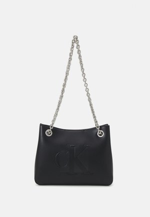 Sac à main en cuir noir avec bandoulières en chaîne argentée et grand logo "CK" embossé sur le devant, sur fond blanc.