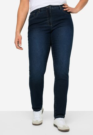 Personne portant un jean slim bleu foncé, des baskets blanches et une chemise blanche, une main sur la hanche et l'autre détendue le long du corps.