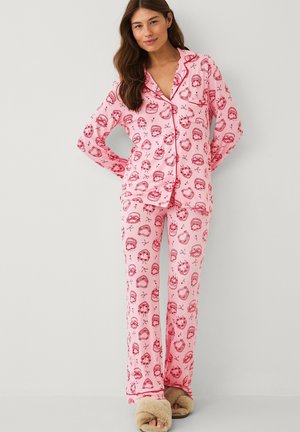 Vrouw draagt een roze pyjama met lange mouwen en knopen, met rode krans- en strikpatronen en beige pluizige pantoffels, staand tegen een effen achtergrond.