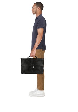 Hombre con polo azul marino, pantalones marrones y zapatillas blancas, sosteniendo un maletín de cuero negro por el asa, de perfil contra un fondo blanco.