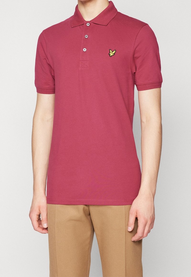 Homme portant un polo à manches courtes bordeaux avec un petit logo d'oiseau jaune sur la poitrine et un pantalon beige.