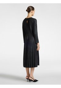 Schwarzes plissiertes Kleid mit langen Ärmeln und einer Schlüsselaugenöffnung am Rücken. Gefertigt aus glattem Material, kombiniert mit schwarzen hohen Schuhen.