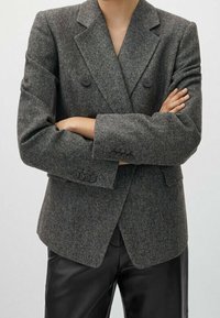 Blazer doppiopetto in tweed grigio testurizzato, con Bottoni neri e rever a notches, abbinato a pantaloni neri su misura.
