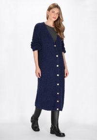 Navyblauer, flauschiger Cardigan mit Knopfleiste, V-Ausschnitt und hochgerollten Ärmeln, kombiniert mit schwarzen Leder-Ankle-Boots.