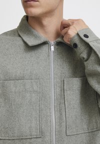 Grijze vilten jas met ritssluiting, twee voorzakken en knoopmanchetten. Voorzien van een klassiek kraagontwerp en een zachte textuur.