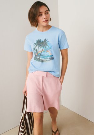 Kvinde iført en lyseblå Maui Hawaii grafisk t-shirt og pink shorts, der holder en stribet netpose og går indendørs på en lys gulvbelægning.