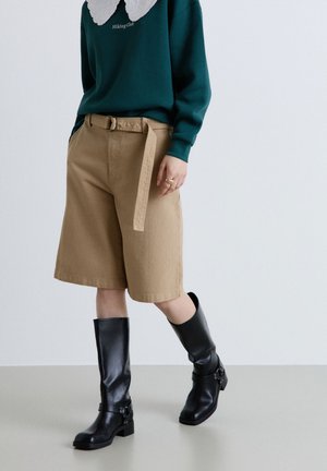 Persona che indossa pantaloncini beige con cintura fino al ginocchio, stivali neri al ginocchio e un maglione verde scuro con colletto di pizzo bianco.